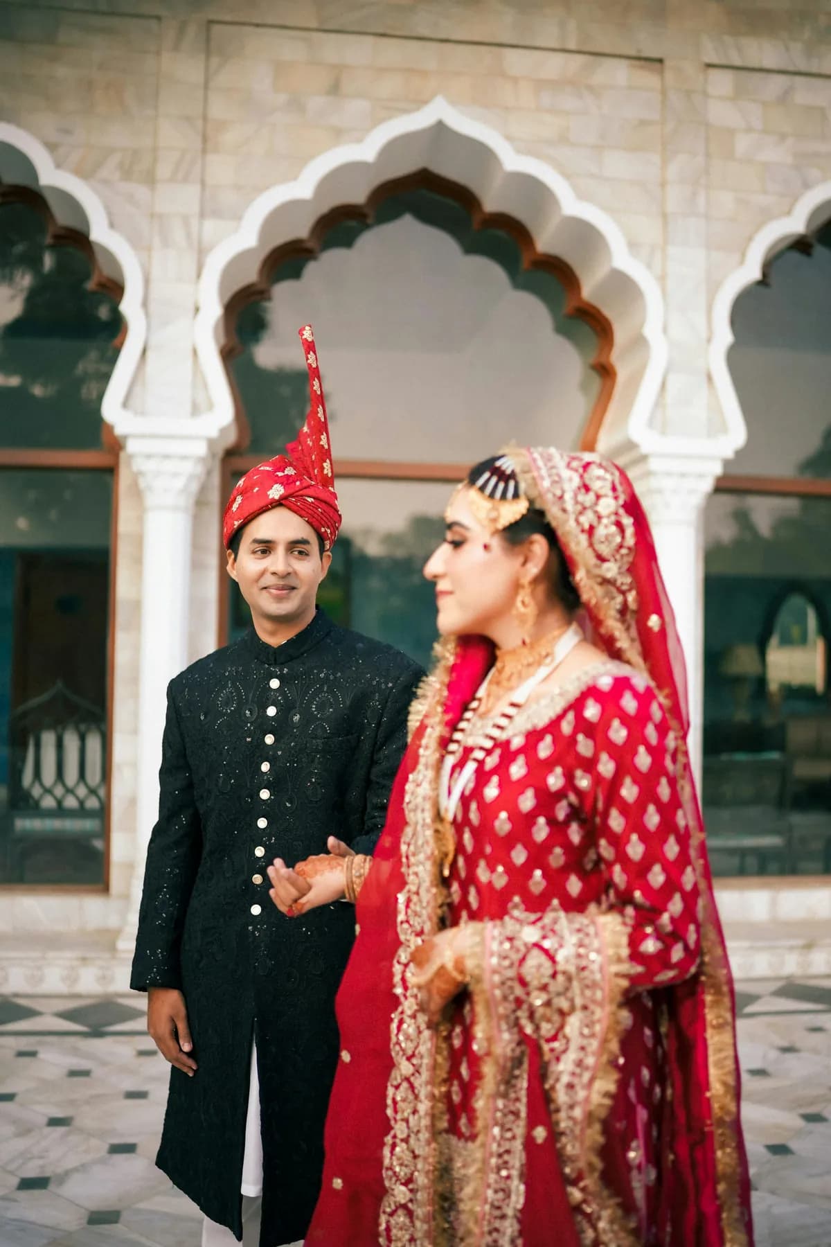 Devika in a red lehenga turning away under white palace arches, Armaan smiling behind her